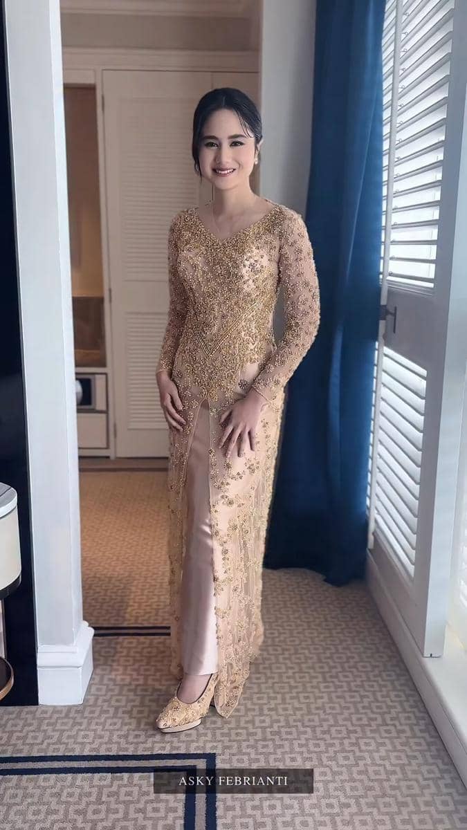 Seorang perempuan mengenakan kebaya modern berwarna emas dengan detail payet, berdiri di dalam ruangan dekat jendela besar.