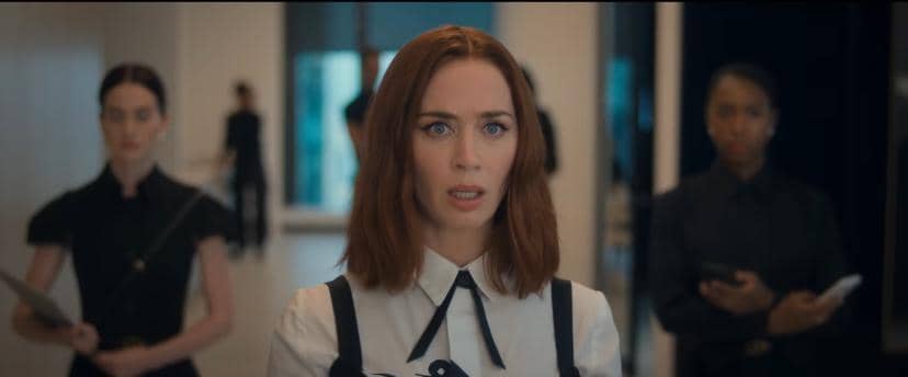 Seorang wanita berambut cokelat mengenakan kemeja putih dengan tali hitam berdiri di koridor kantor, dengan dua wanita lain di latar belakang.