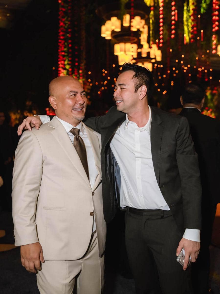 Dua pria mengenakan setelan jas berdiri berdampingan sambil tersenyum di acara malam dengan pencahayaan hangat dan dekorasi lampu gantung.