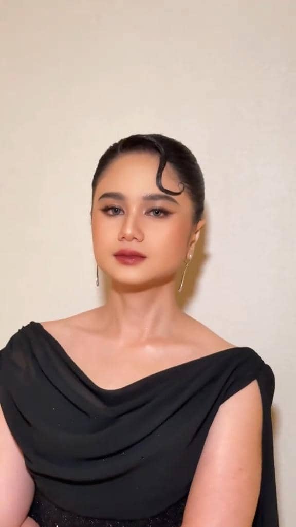 Seorang perempuan bergaun hitam elegan dengan riasan wajah glamor dan anting panjang berpose di depan dinding berwarna krem.