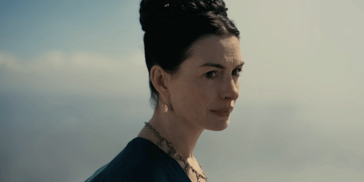 Seorang wanita berambut gelap dengan gaya sanggul mengenakan anting dan kalung, berdiri di luar ruangan dengan latar langit cerah.