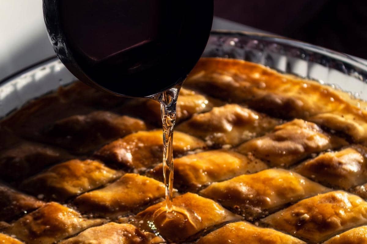 Sirup kental dituangkan ke atas potongan baklava berwarna keemasan yang baru dipanggang dalam wadah kaca besar.