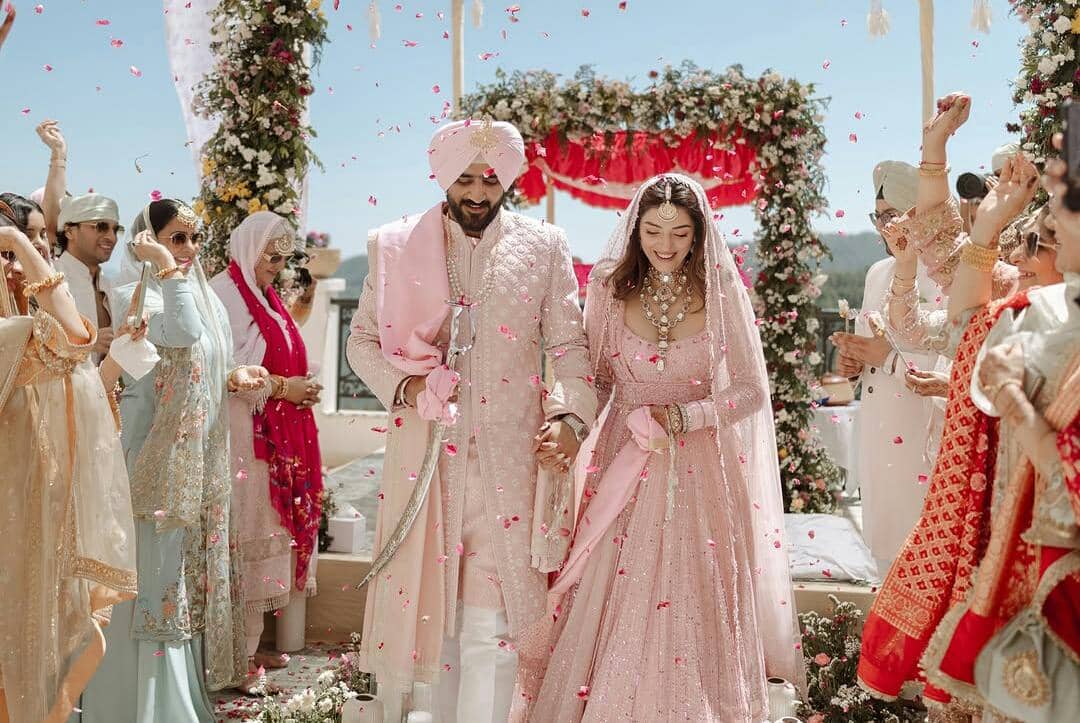 Pasangan pengantin mengenakan busana tradisional India berwarna merah muda berjalan di bawah taburan bunga dalam upacara pernikahan outdoor.