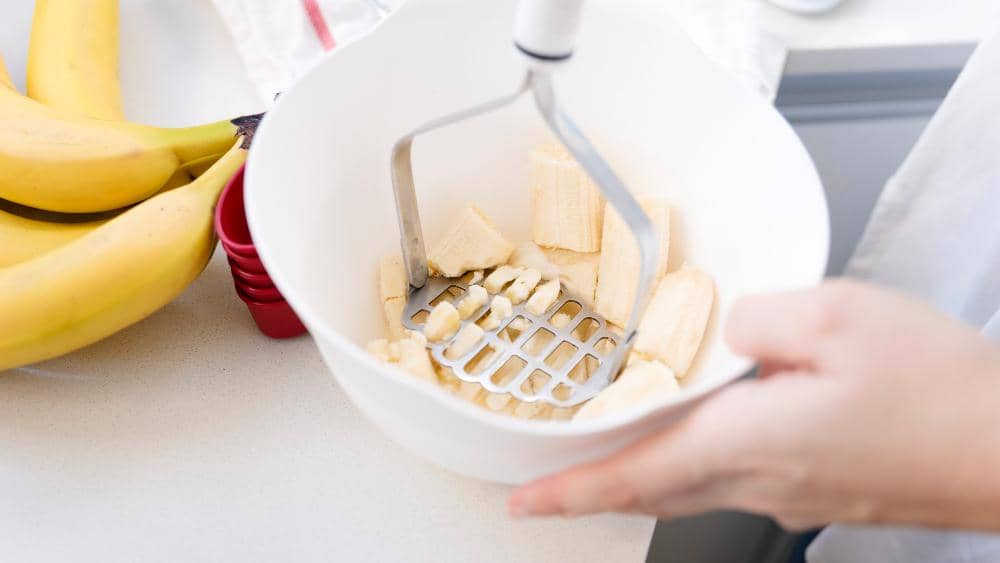 Tangan sedang menghaluskan potongan pisang matang dalam mangkuk putih menggunakan alat penumbuk di dapur.