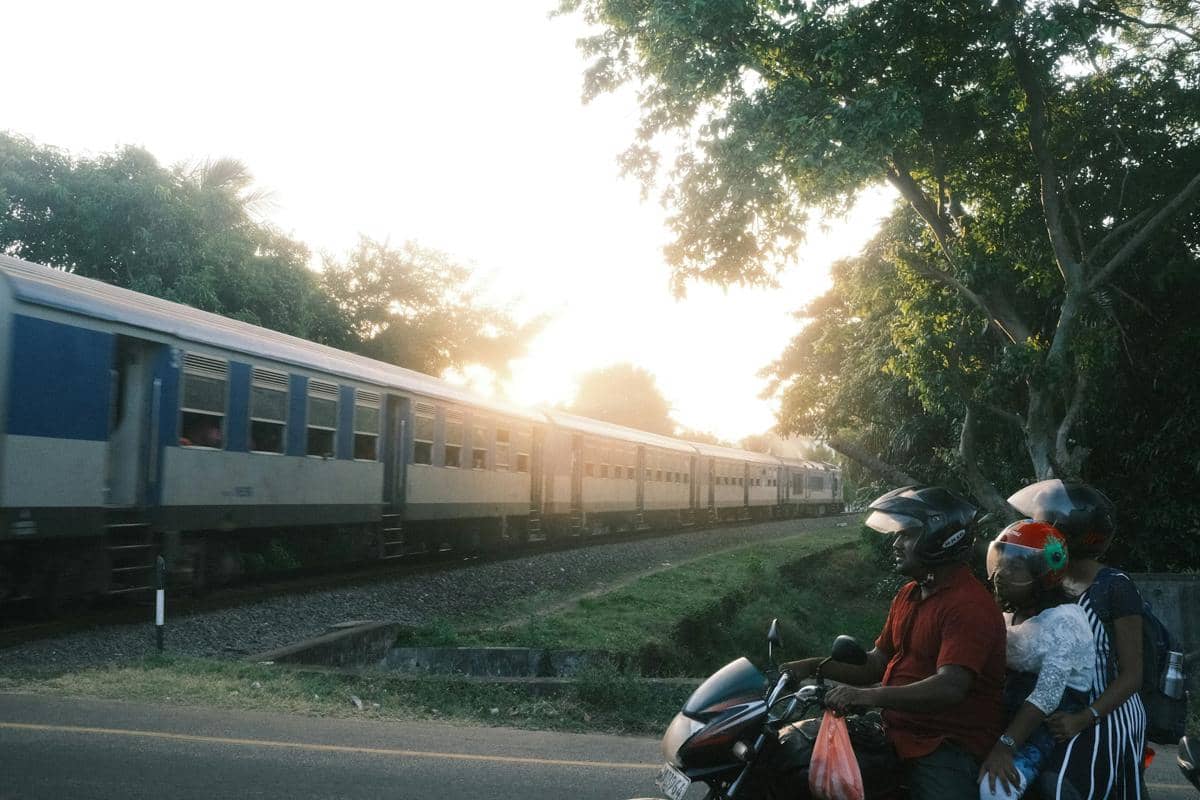 Tiga orang berboncengan di sepeda motor berhenti di tepi jalan saat kereta api melintas di rel di bawah sinar matahari sore.