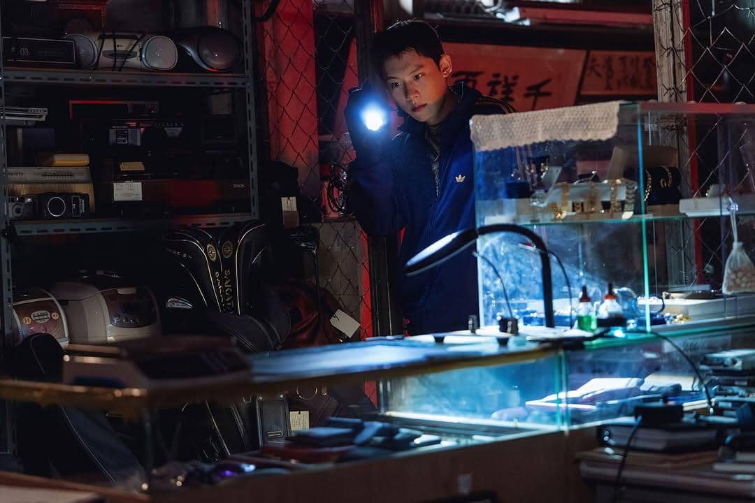 Seorang pria mengenakan jaket biru memegang senter di ruangan gelap penuh barang elektronik dan rak kaca berisi peralatan kecil.