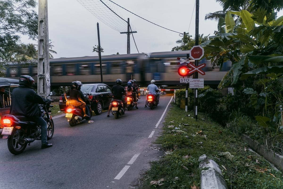 Beberapa pengendara motor dan mobil berhenti di perlintasan kereta api dengan lampu merah menyala saat kereta melintas di sore hari.