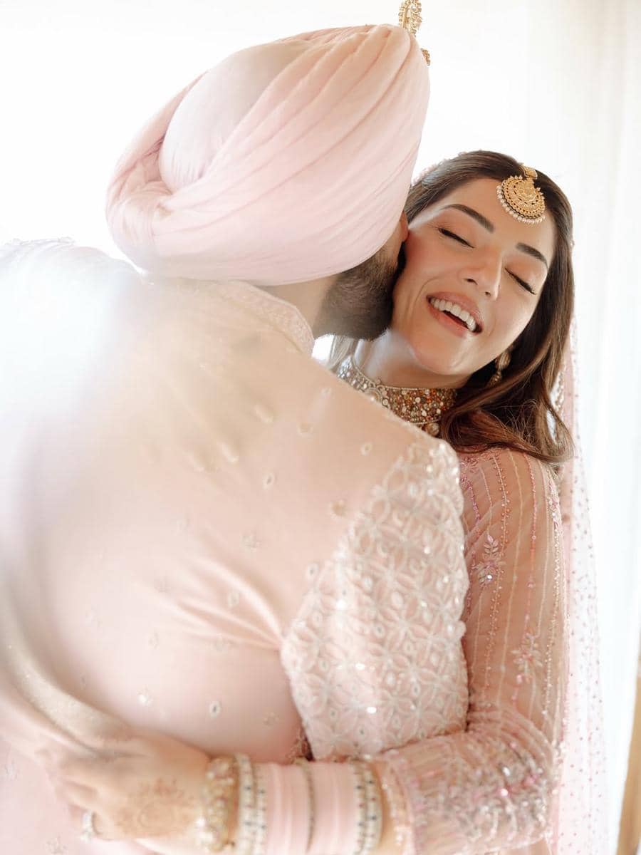 Pasangan pengantin berpakaian tradisional India berwarna pastel saling berpelukan dengan ekspresi bahagia di hari pernikahan.