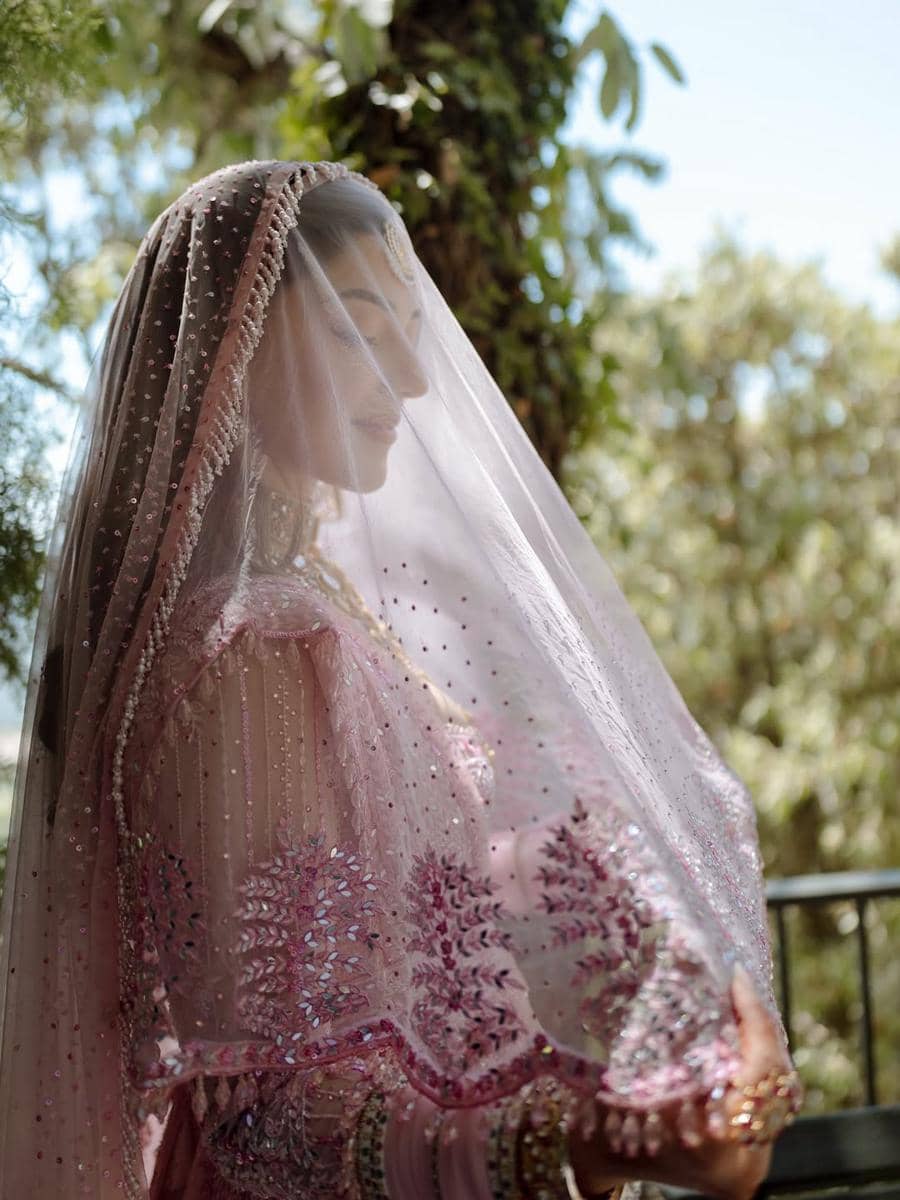Seorang pengantin mengenakan gaun tradisional berwarna merah muda dengan kerudung berhias manik-manik di luar ruangan yang rindang.