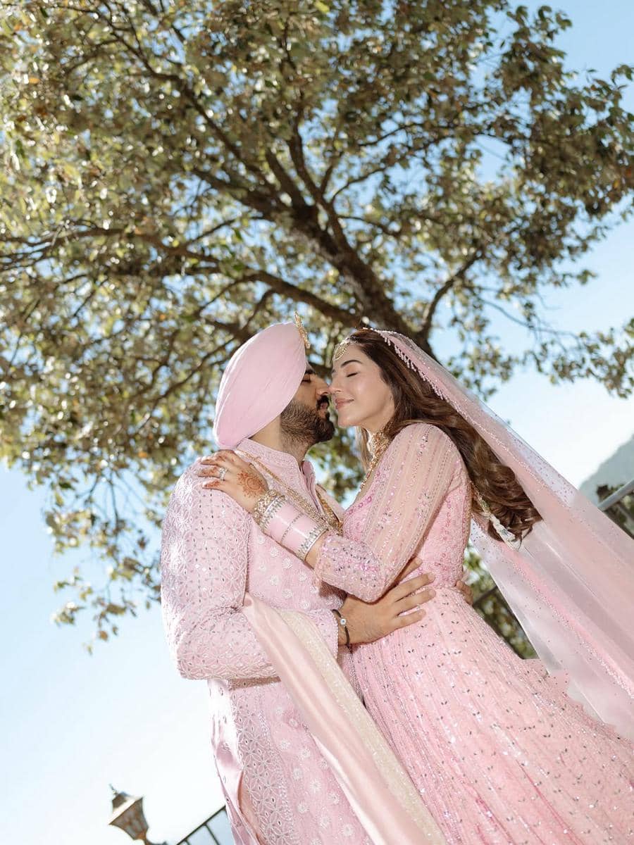 Pasangan pengantin mengenakan busana tradisional India berwarna merah muda saling berpelukan di bawah pohon pada hari pernikahan.