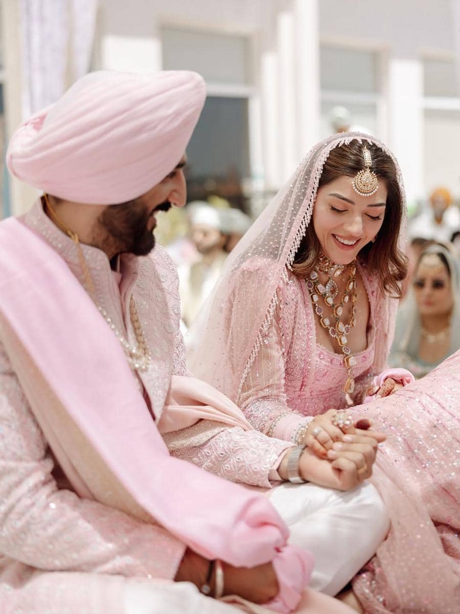 Pasangan pengantin mengenakan busana tradisional India berwarna merah muda lembut sedang tersenyum dan saling menggenggam tangan di upacara pernikahan.