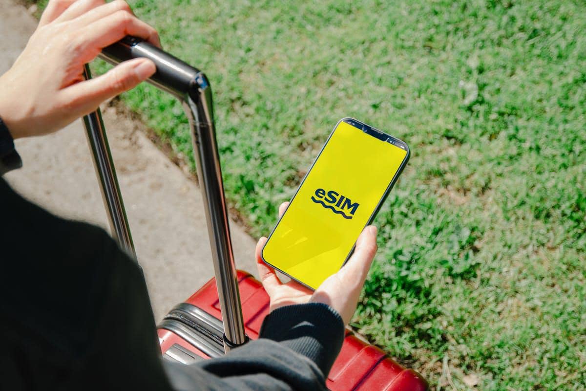 Seseorang memegang ponsel dengan layar bertuliskan eSIM berwarna kuning sambil menarik koper merah di area luar ruangan.