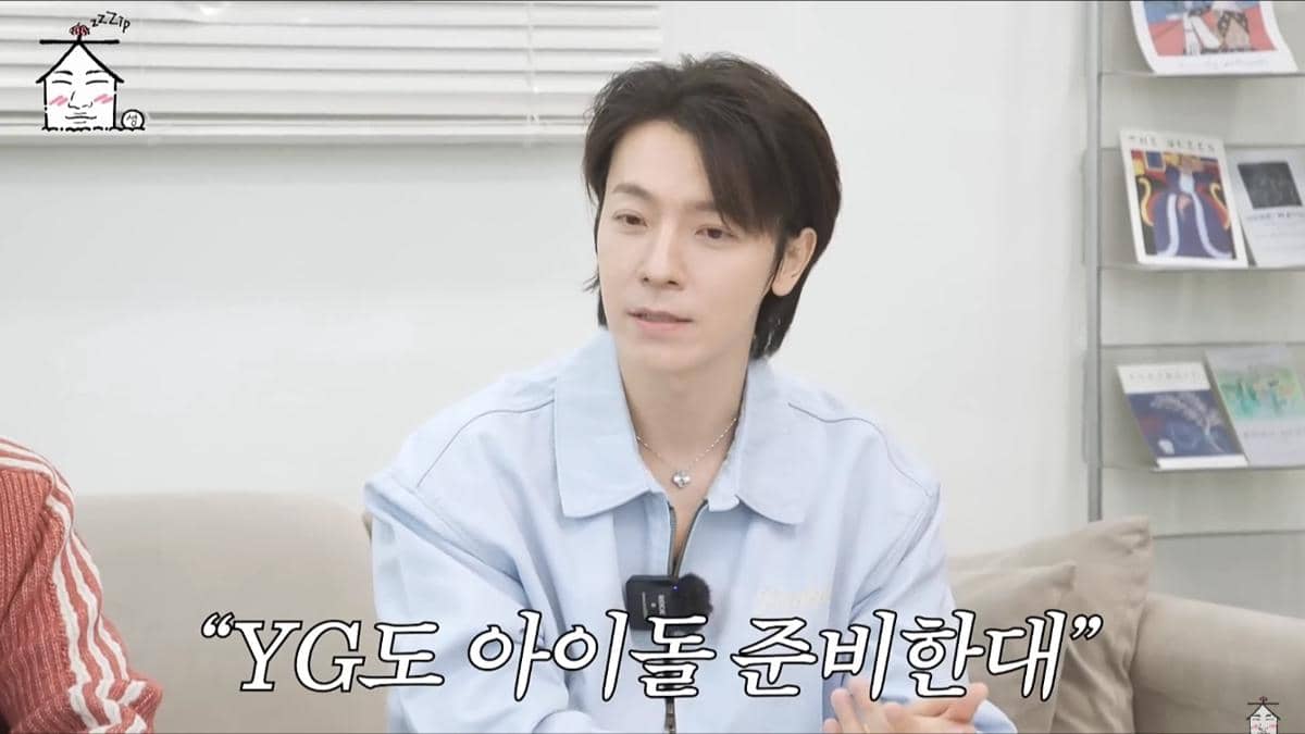 Seorang pria berambut hitam mengenakan kemeja biru muda sedang berbicara di ruang santai dengan latar belakang rak buku dan tulisan Korea di layar.