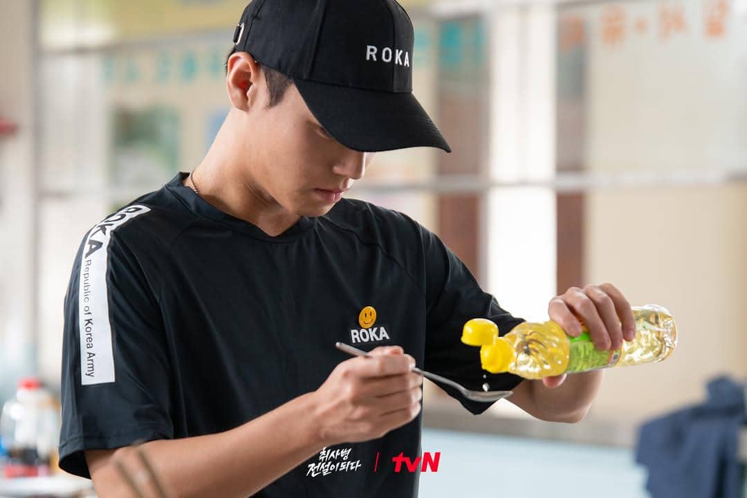 Seorang pria mengenakan topi dan kaus bertuliskan ROKA sedang menuangkan minyak ke sendok di dapur dalam adegan drama The Legend of Kitchen Soldier.