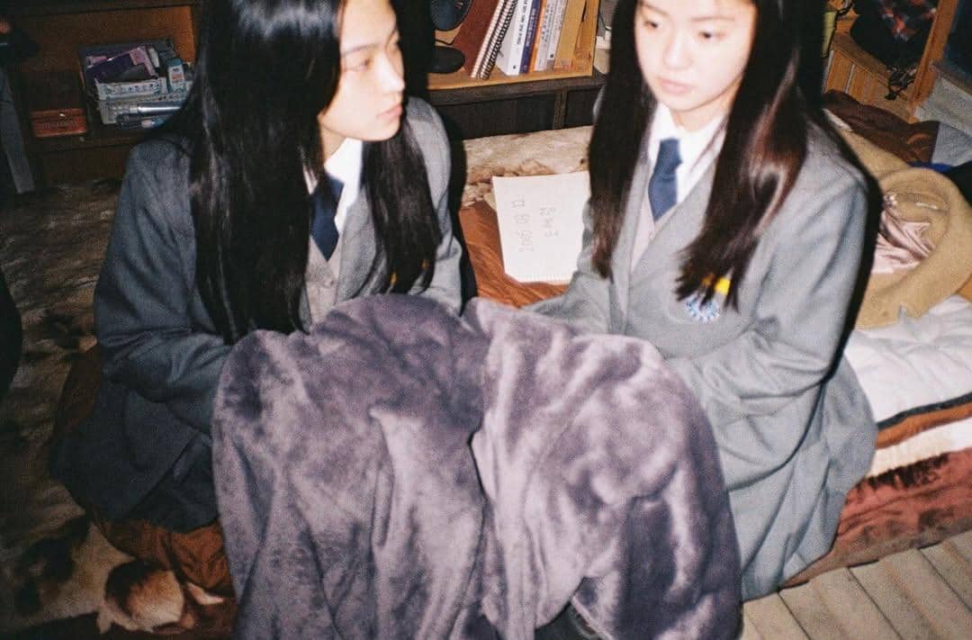 Dua remaja berseragam sekolah duduk di kamar dengan selimut abu-abu di pangkuan, tampak berbincang serius di suasana hangat.