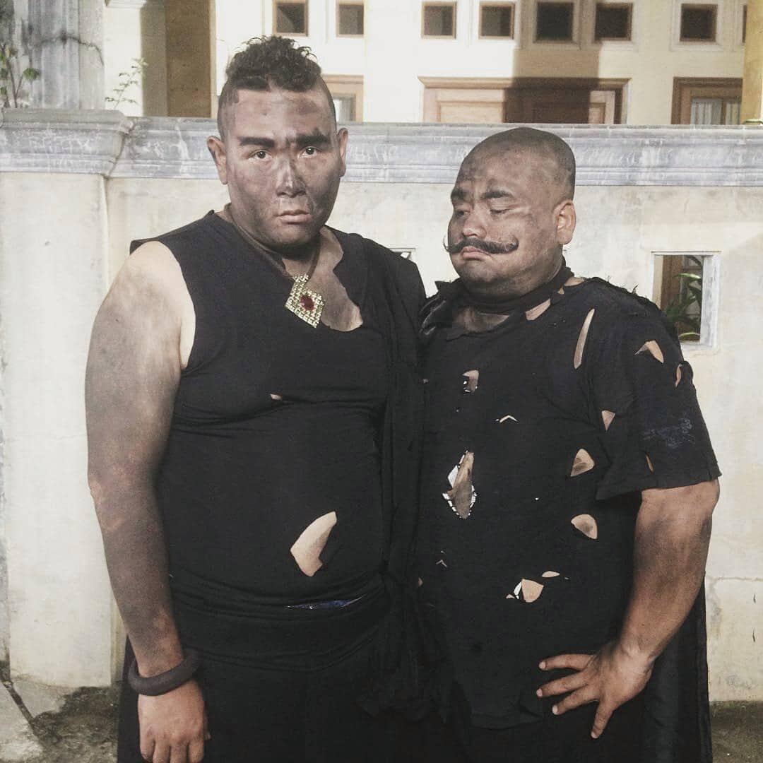 Dua pria mengenakan pakaian hitam robek dan wajah berlumur jelaga berdiri berdampingan di depan bangunan bercat terang.