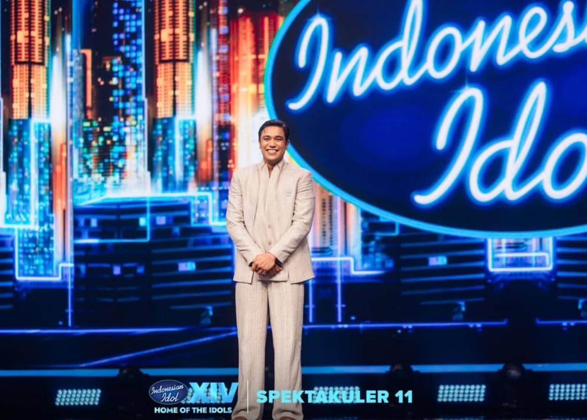 Seorang kontestan berdiri di panggung Indonesian Idol musim ke-14 dengan latar belakang logo besar dan pencahayaan warna biru cerah.