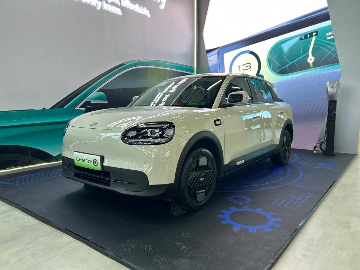 Mobil listrik Chery Q berwarna putih dipamerkan di sebuah stan pameran otomotif dengan latar belakang layar digital besar.