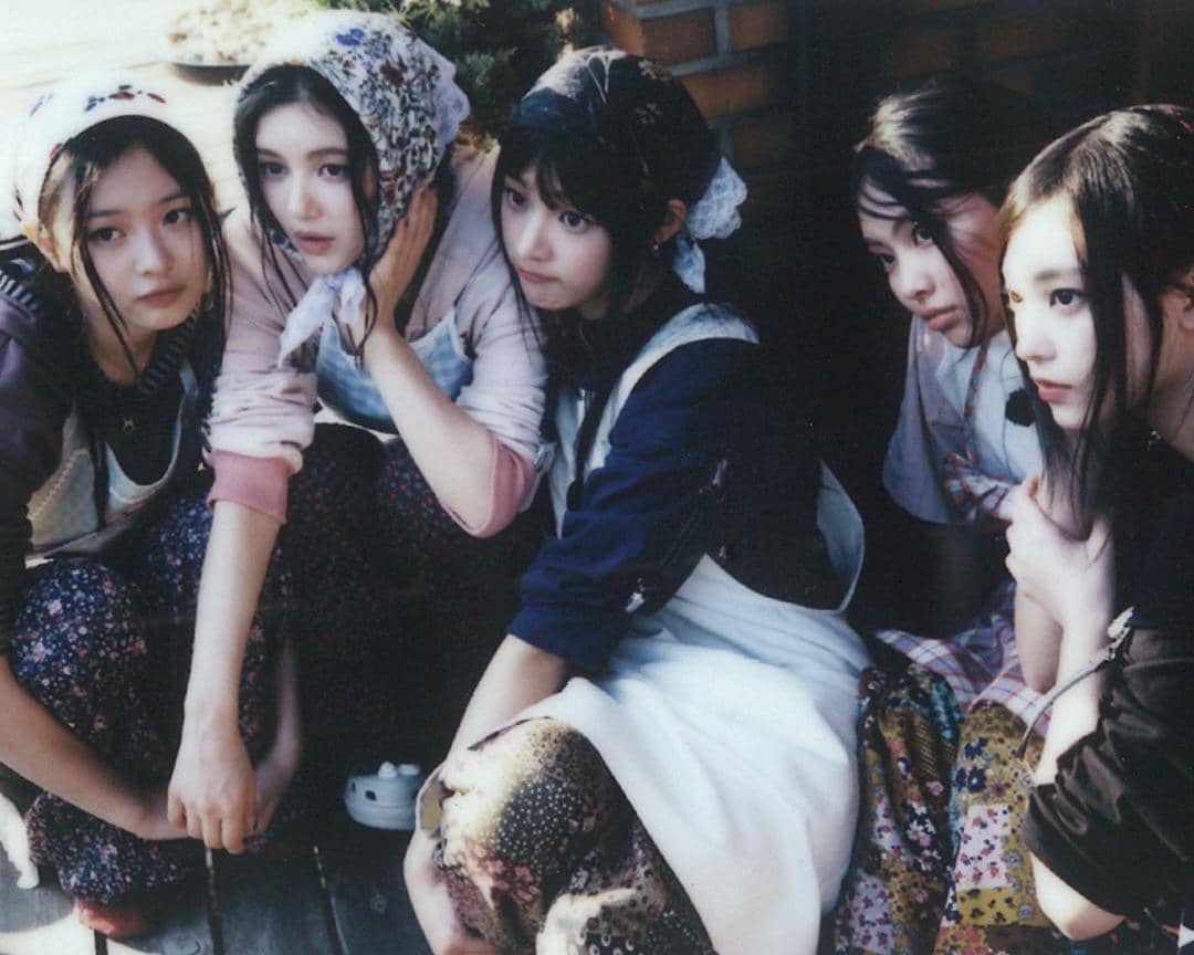 Lima perempuan muda duduk berdekatan mengenakan pakaian bergaya retro dengan penutup kepala bermotif bunga di suasana luar ruangan.