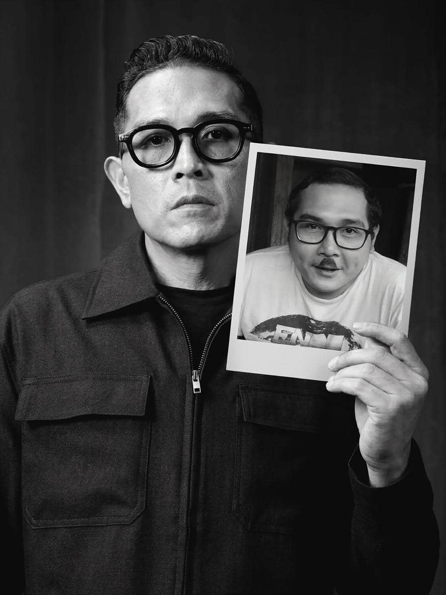 Seorang pria berkacamata mengenakan jaket hitam memegang foto dirinya di masa lalu dengan penampilan berbeda dalam potret hitam putih.