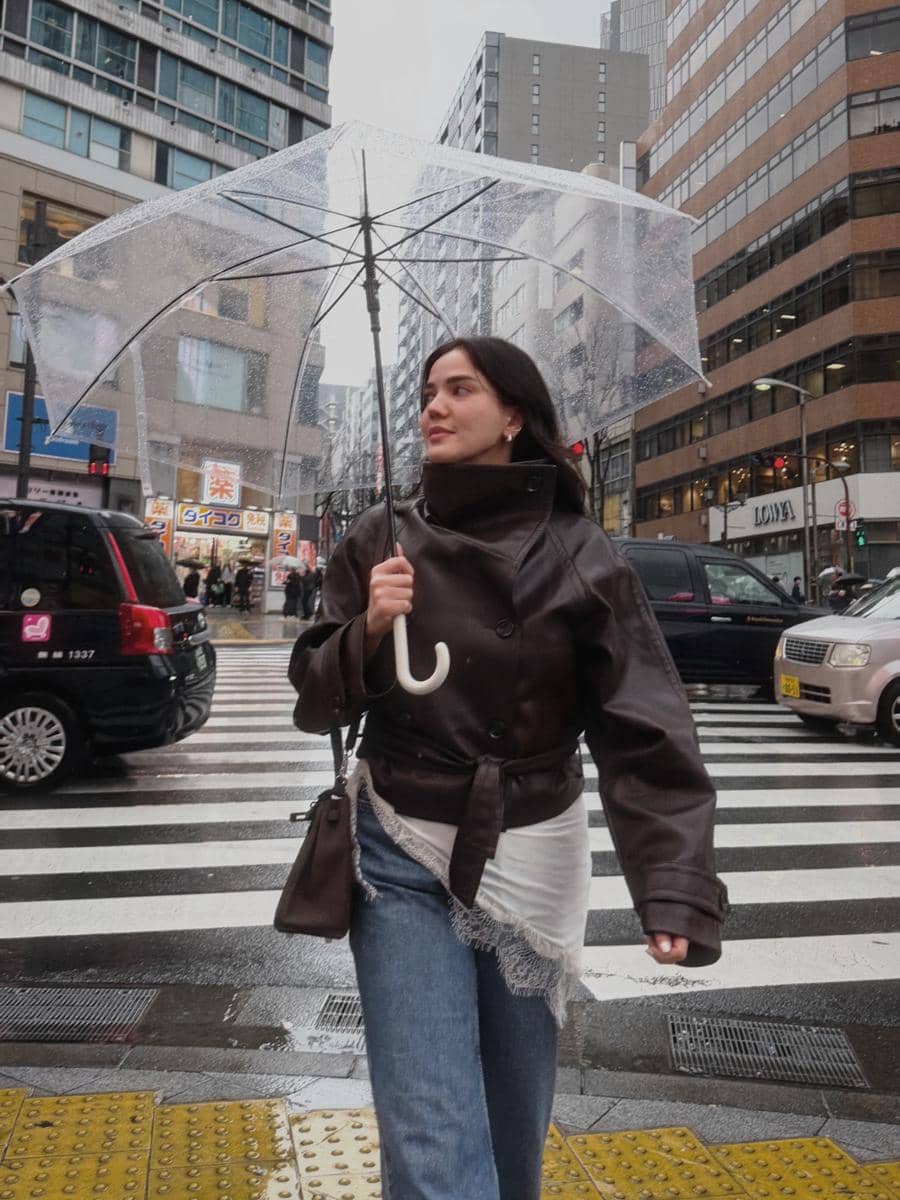 Seorang wanita berjalan di penyeberangan jalan kota besar sambil memegang payung transparan di bawah cuaca hujan.