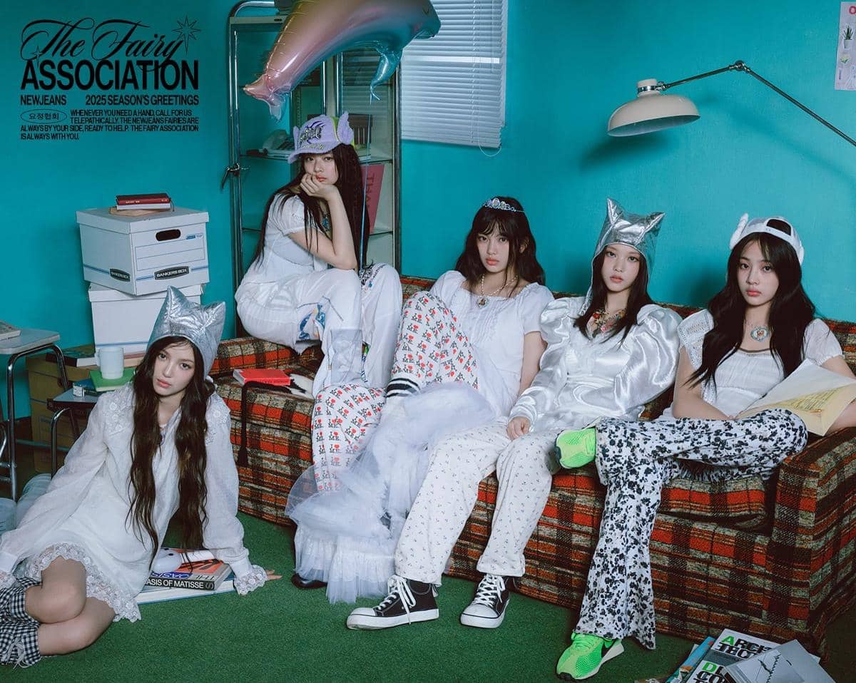 Lima anggota grup musik duduk di sofa bergaya retro dengan busana putih dan aksesori unik di ruangan berwarna biru kehijauan.