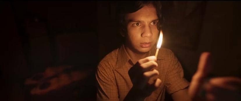 Seorang pria muda memegang korek api yang menyala di ruangan gelap dengan ekspresi tegang, diterangi cahaya api kecil di depannya.
