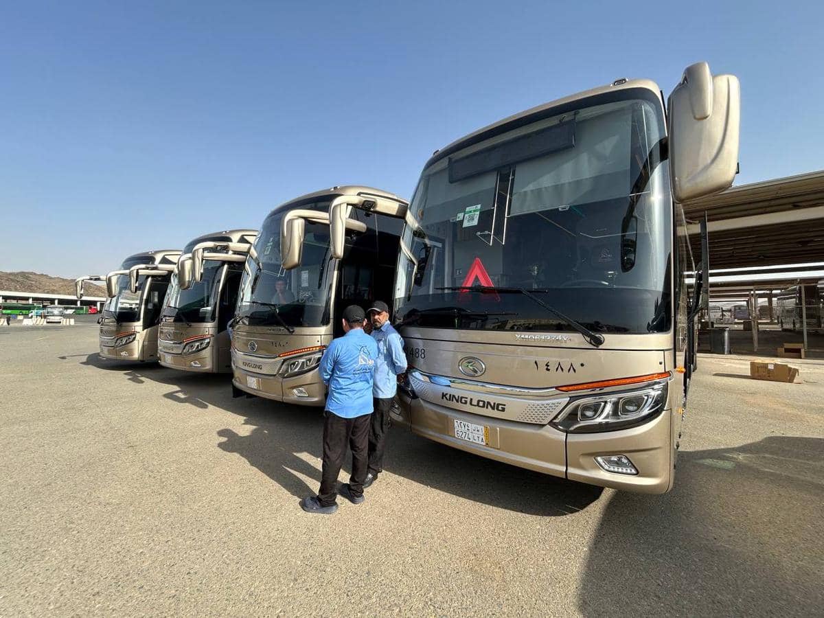 Deretan bus berwarna krem bertuliskan King Long terparkir di area terbuka dengan dua petugas mengenakan seragam biru berbincang di depannya.