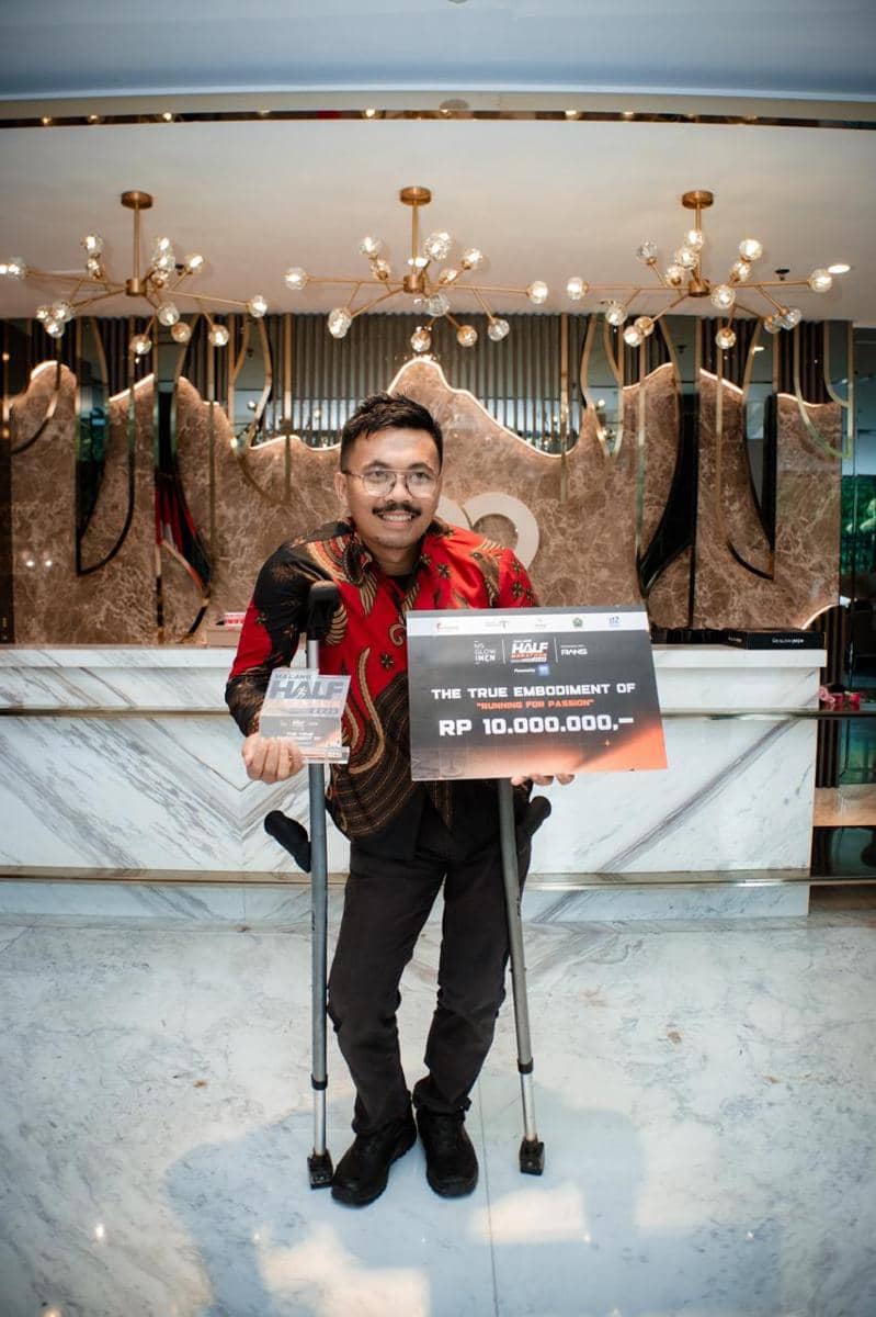 Seorang pria berdiri dengan tongkat penyangga sambil memegang papan hadiah senilai Rp10.000.000 di acara Malang Half Marathon 2026.