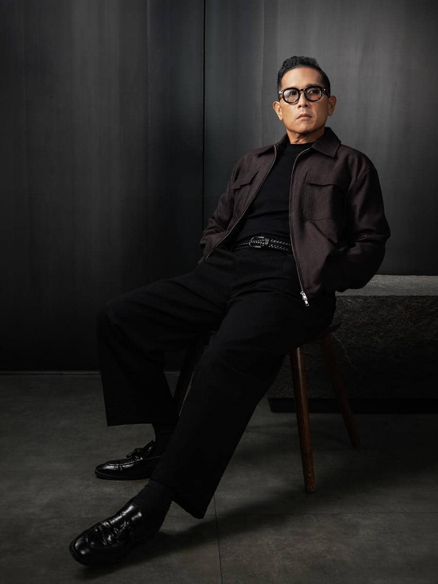 Seorang pria mengenakan pakaian serba hitam duduk di kursi kayu dengan latar belakang dinding gelap bergaya modern minimalis.
