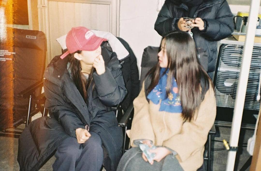 Dua perempuan duduk berdampingan mengenakan pakaian hangat di lokasi syuting, berbincang santai di kursi lipat.