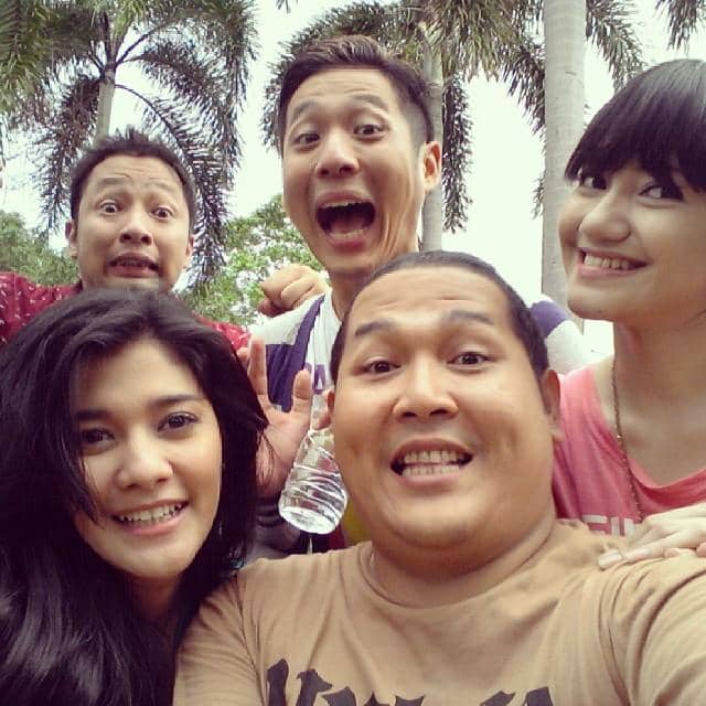 Lima orang berpose ceria bersama di luar ruangan dengan latar belakang pohon palem dan suasana santai penuh tawa.