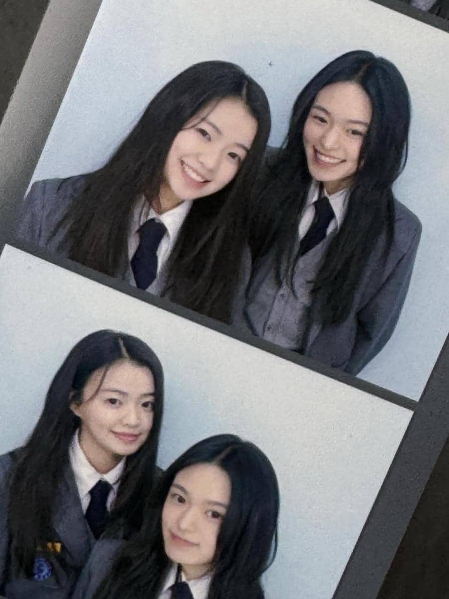 Dua remaja perempuan mengenakan seragam sekolah abu-abu dengan dasi hitam berpose tersenyum di depan latar polos.