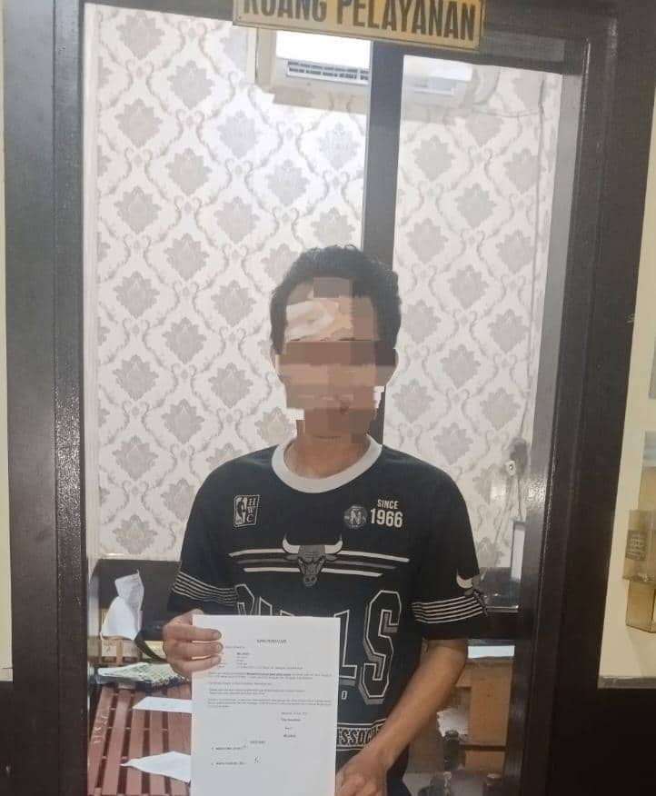 Seorang pria berdiri di dalam ruangan pelayanan kepolisian memegang selembar dokumen dengan wajah yang sebagian diburamkan.