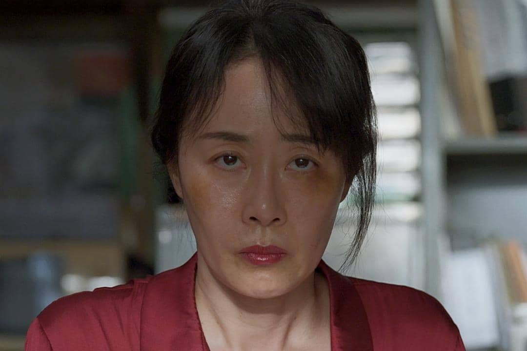 Seorang wanita berwajah serius mengenakan pakaian merah dalam adegan drama Korea Gold Land, dengan latar ruangan redup.
