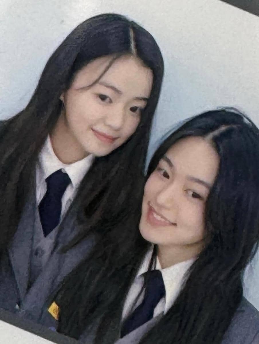 Dua perempuan berambut panjang mengenakan seragam sekolah abu-abu dengan dasi biru gelap berpose sambil tersenyum di depan kamera.