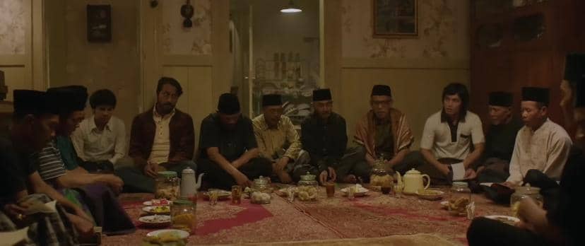 Sekelompok pria duduk melingkar di ruang tamu dengan karpet merah, mengenakan peci dan pakaian sopan, tampak sedang mengadakan doa bersama.