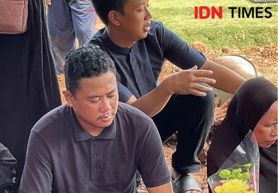 Seorang pria mengenakan pakaian hitam duduk dengan wajah sedih di area pemakaman bersama beberapa orang lainnya.