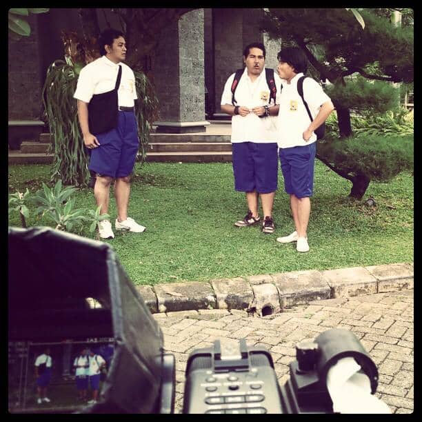 Tiga pria mengenakan seragam sekolah menengah berdiri di halaman hijau dengan kamera film di latar depan.