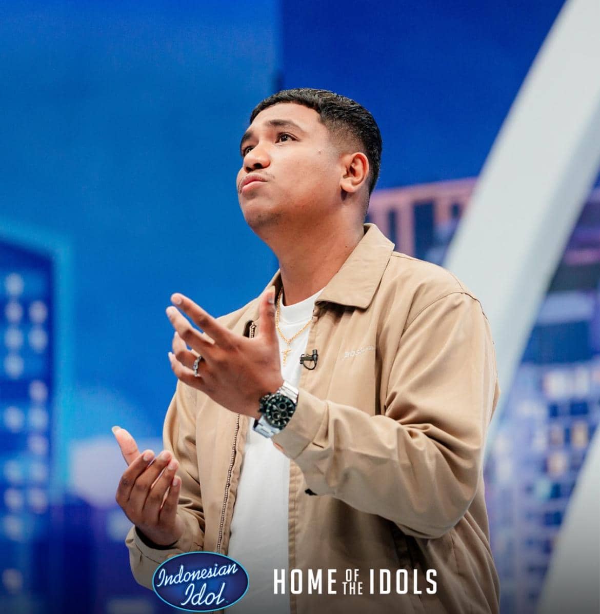 Seorang peserta Indonesian Idol mengenakan jaket krem dan kaus putih sedang bernyanyi di atas panggung dengan latar biru bertuliskan Home of the Idols.