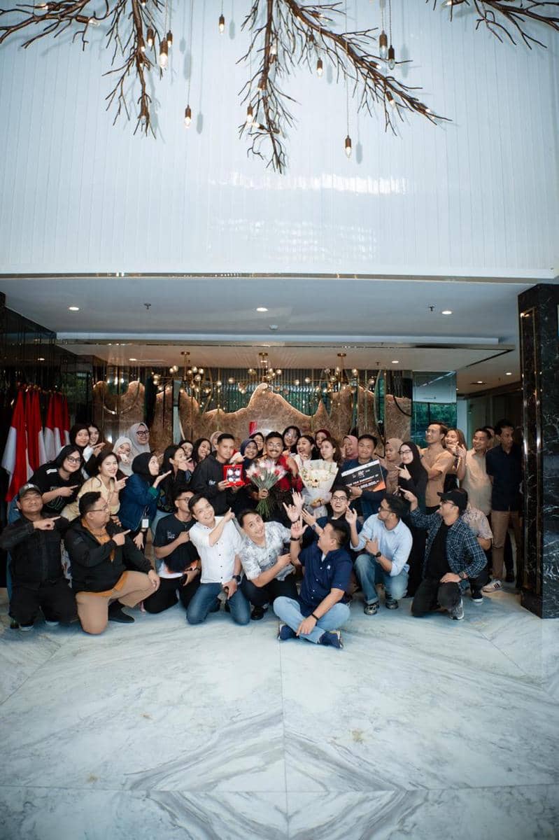 Sekelompok orang berpose bersama di dalam ruangan mewah dengan pencahayaan gantung artistik, beberapa memegang buket bunga dan papan hadiah.