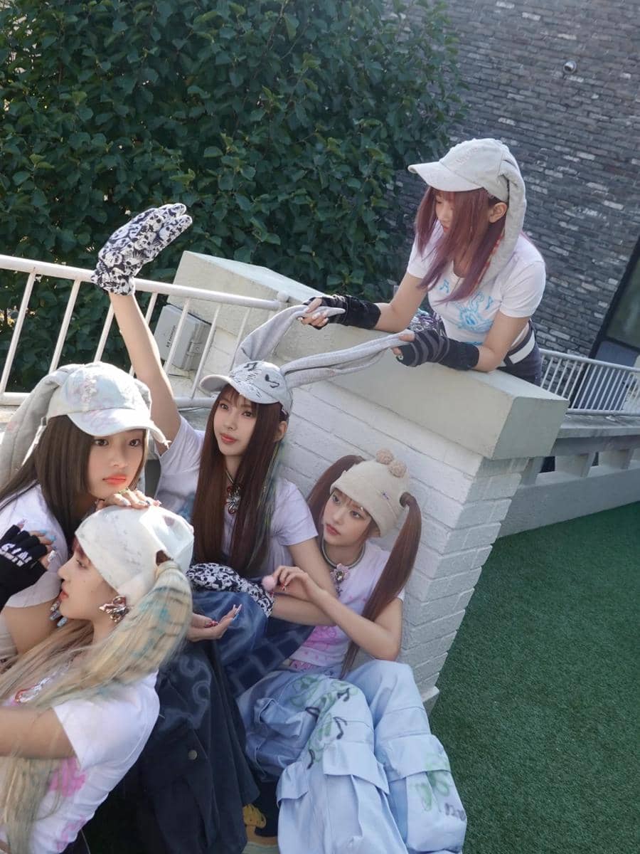 Lima perempuan muda mengenakan topi dan pakaian kasual berwarna cerah sedang berpose bersama di area luar dengan latar dinding bata putih dan tanaman hijau.