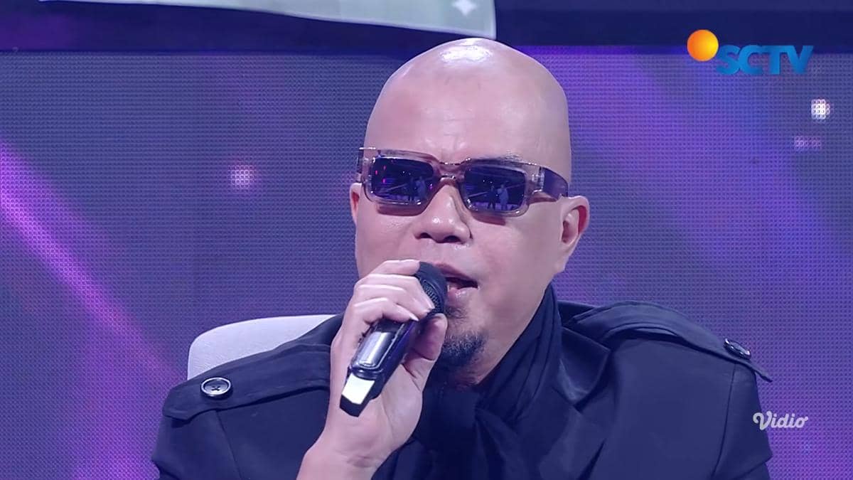 Seorang pria berkepala plontos mengenakan kacamata hitam dan jaket hitam sedang memegang mikrofon di atas panggung dengan latar ungu.
