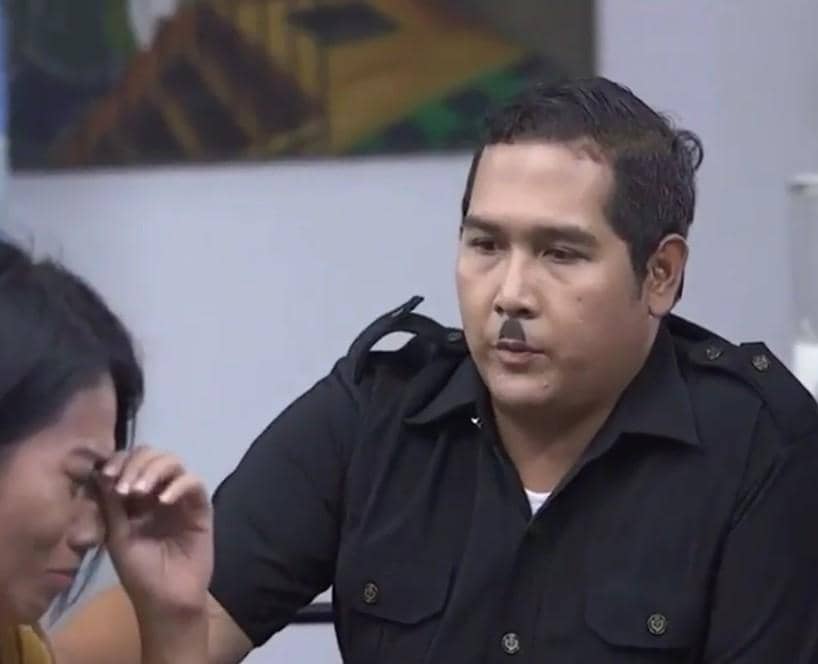 Seorang pria berseragam hitam berbicara dengan seorang wanita yang tampak sedih di dalam ruangan dengan latar dinding putih.