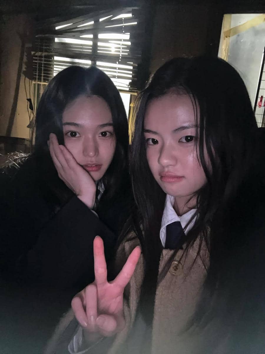 Dua perempuan muda berpose bersama di ruangan redup, salah satunya mengangkat tangan membentuk tanda V sambil tersenyum tipis.