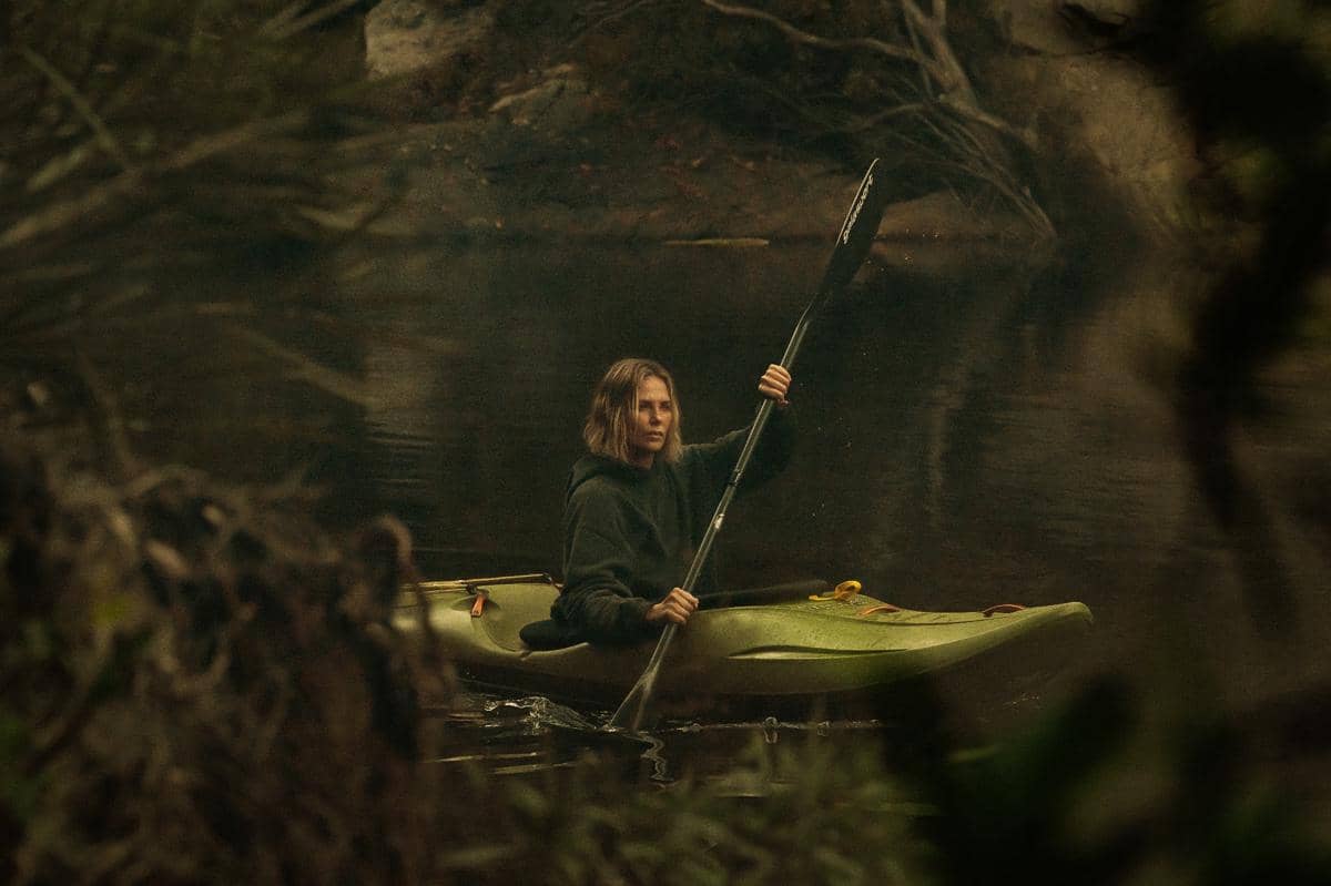 Seorang perempuan mendayung kayak hijau di sungai tenang yang dikelilingi pepohonan lebat dalam suasana gelap dan misterius.