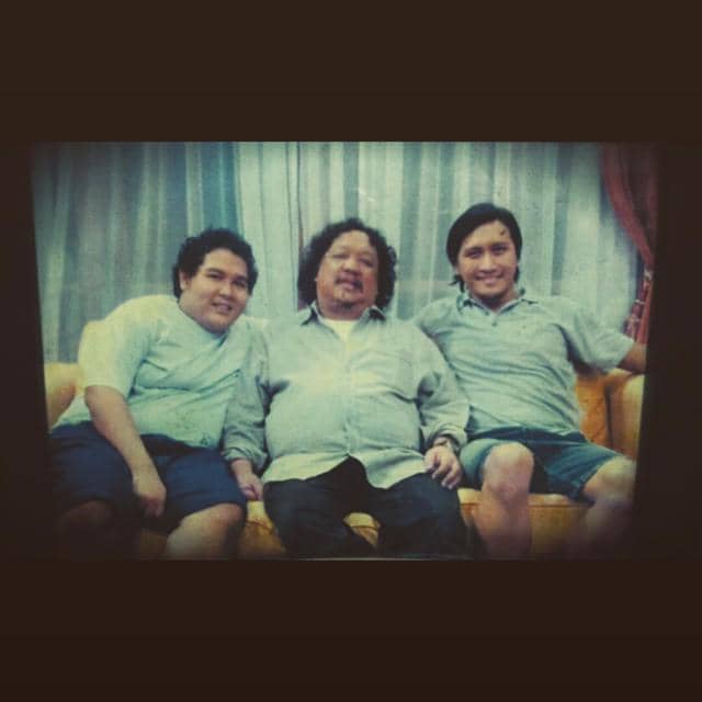 Tiga pria duduk di sofa berwarna kuning di ruang tamu dengan tirai abu-abu di latar belakang, semuanya mengenakan kemeja kasual.