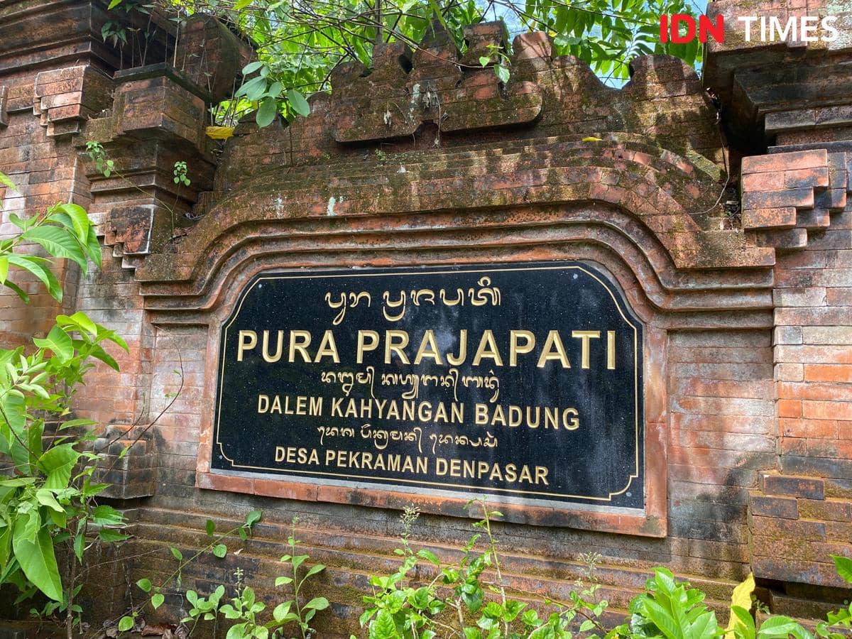 Papan nama batu bata merah bertuliskan Pura Prajapati Dalem Kahyangan Badung di Desa Pekraman Denpasar, dikelilingi tanaman hijau.