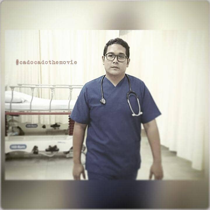 Seorang pria mengenakan seragam medis biru dan stetoskop di lehernya berdiri di ruang rumah sakit dengan tirai putih di latar belakang.