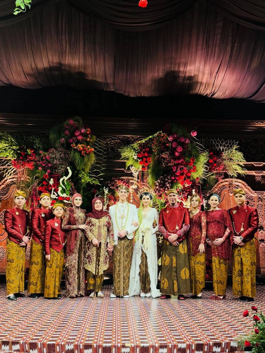 Keluarga besar mengenakan busana adat Jawa berwarna merah dan cokelat berpose bersama pengantin di pelaminan berhias bunga merah dan hijau.